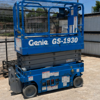  LiftsBlue Genie GS-1930 scissor lift parked on a concrete surface, surrounded by fencing and industrial buildings. The lift is in a lowered position.