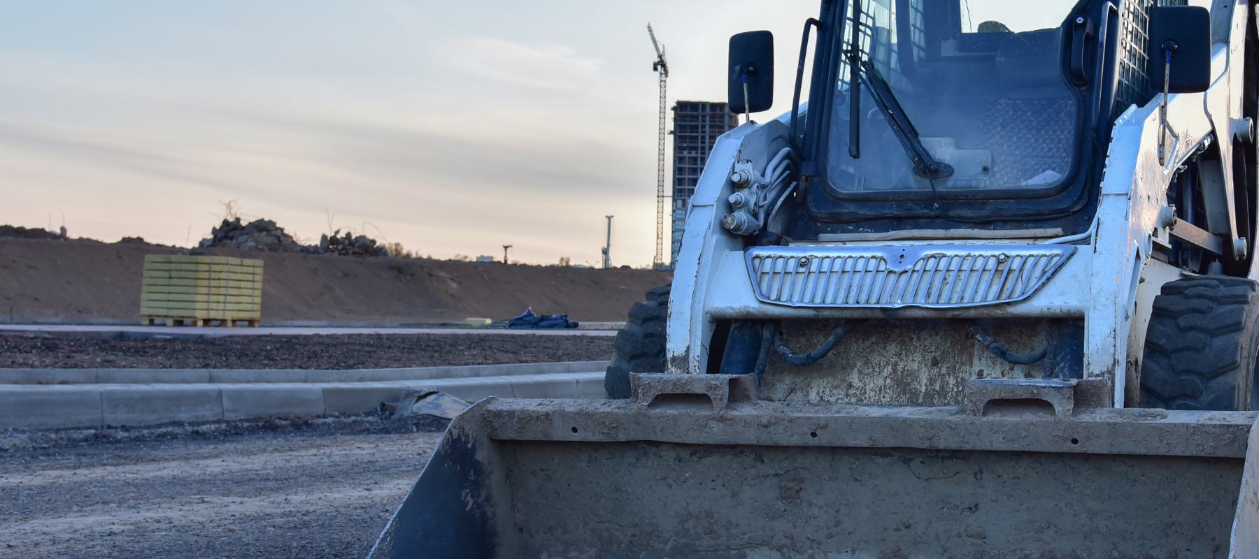 A large construction vehicle with a front bucket on a dirt site. Behind it, mounds of earth and a tall crane tower against a cloudy sky.